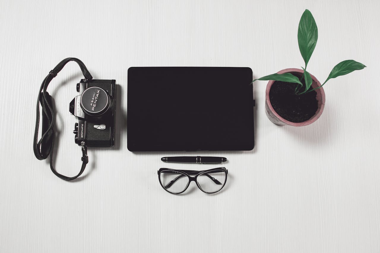 An overhead view of a neat desk setup featuring a camera, tablet, glasses, pen, and plant, emphasizing modern work aesthetics.